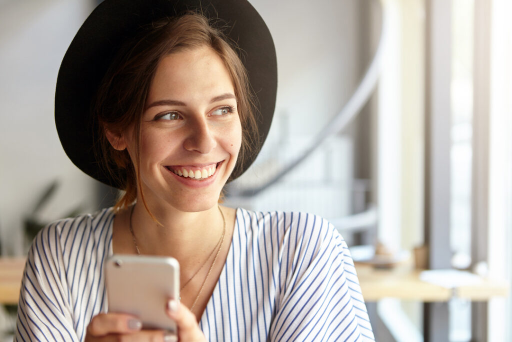 Indoor shot of happy carefree young woman with cute smile enjoying free wi-fi at coffee shop, surfing internet on electronic gadget, messaging friends online. People, technology and communication