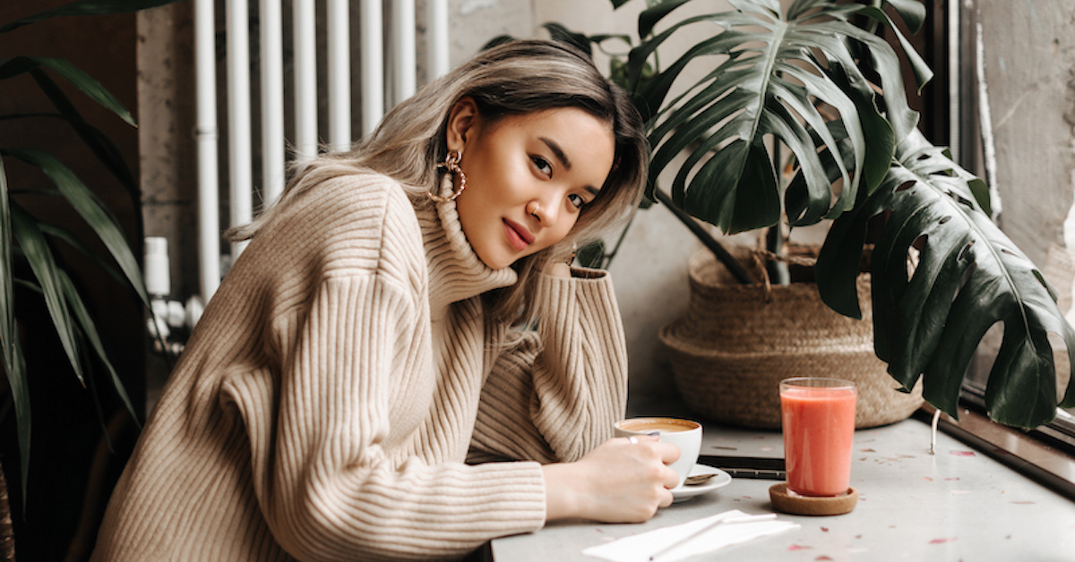 Blonde Asian girl in beige oversized sweater sits in cafe with cup of coffee and carrot juice