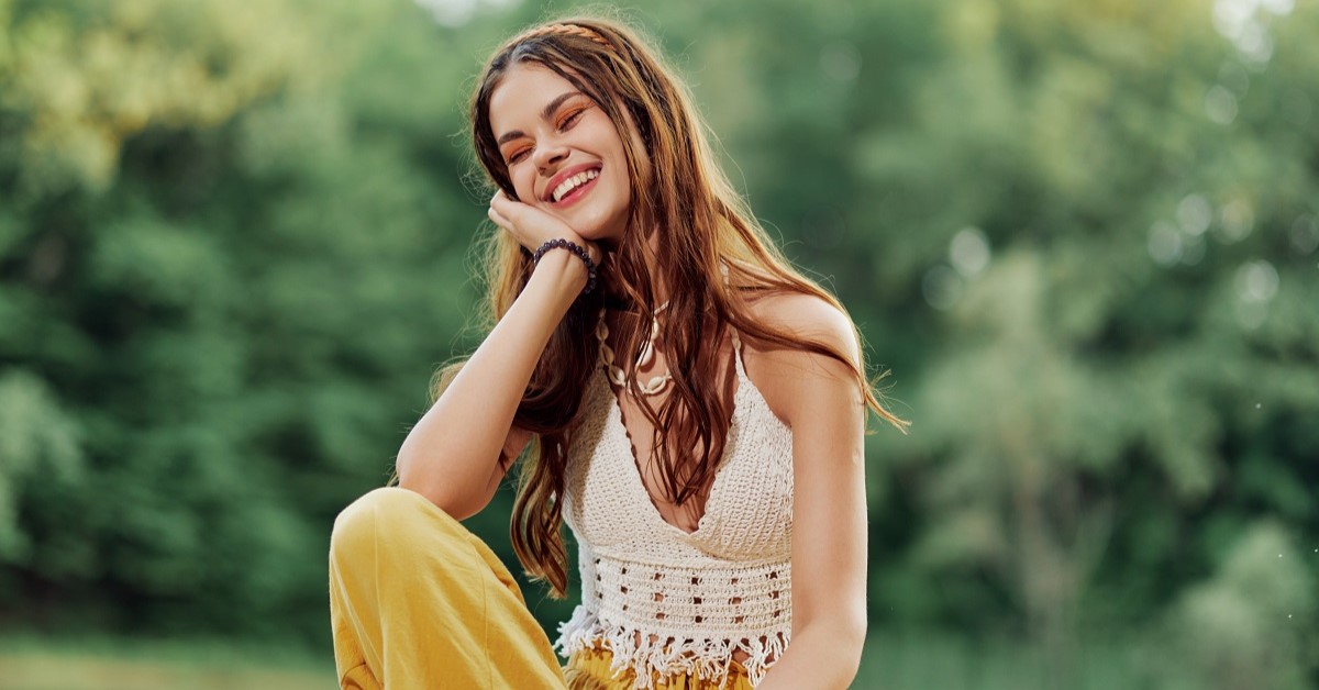 A young woman smiling in an image of a hippie and eco-clothes sitting outdoors on a bridge by a lake wearing a hat and yellow pants in the summer sunset