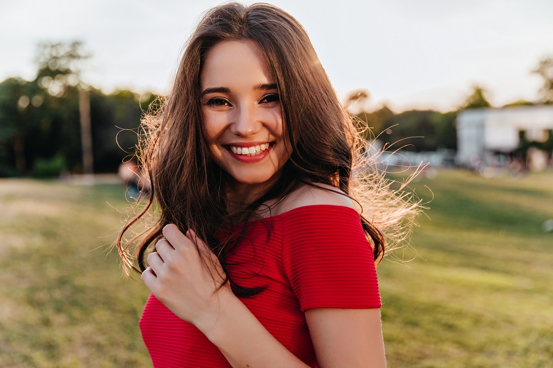 Portrait of blithesome caucasian woman with dark hair standing on blur nature background. Glad brunette girl in red dress smiling to camera during rest in park.