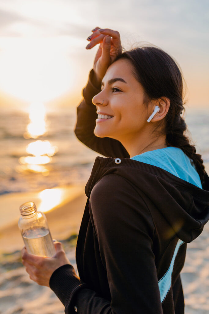 Attractive slim woman doing sports exercises on the beach at sunrise, wearing sports attire, drinking water from a bottle, listening to music with wireless earphones on a hot summer day.