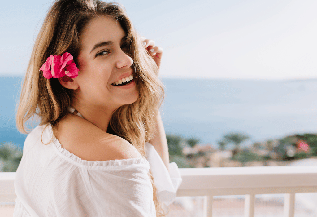 Beautiful smiling woman with a flower on her ear wearing a white top with the sea as her background