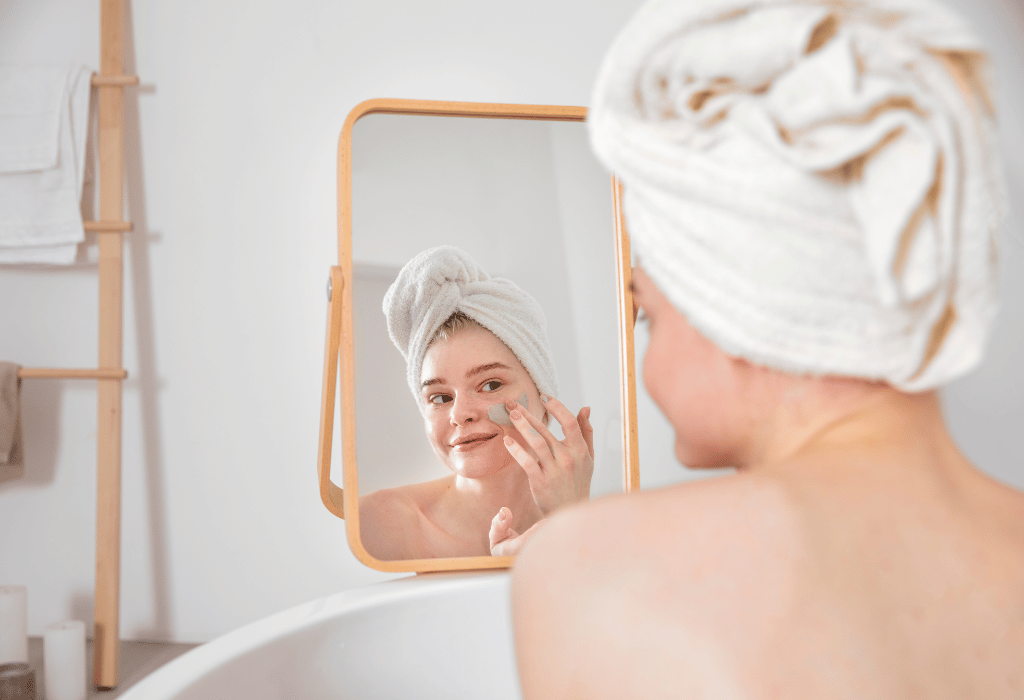 Woman after a bath wearing a towel on her head and putting a clay mask on her face while looking at the mirror