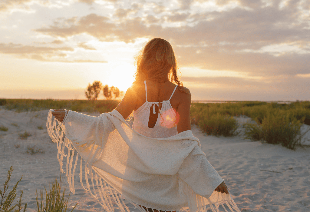 Woman in a white mini dress and shawl facing the sunset with her back on the camera near the beach